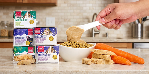 Image of a can of Open Farm wet cat food, a red container of wet cat food and a tin can of sardines for cats on a white countertop with a white bowl filled with wet cat food